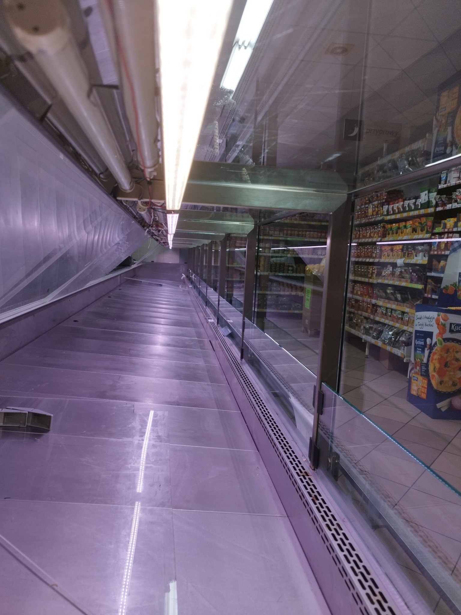 Wnętrze chłodni sklepowej z zamontowanym oświetleniem liniowym LED pod sufitem, widoczne półki i szklane przegrody.