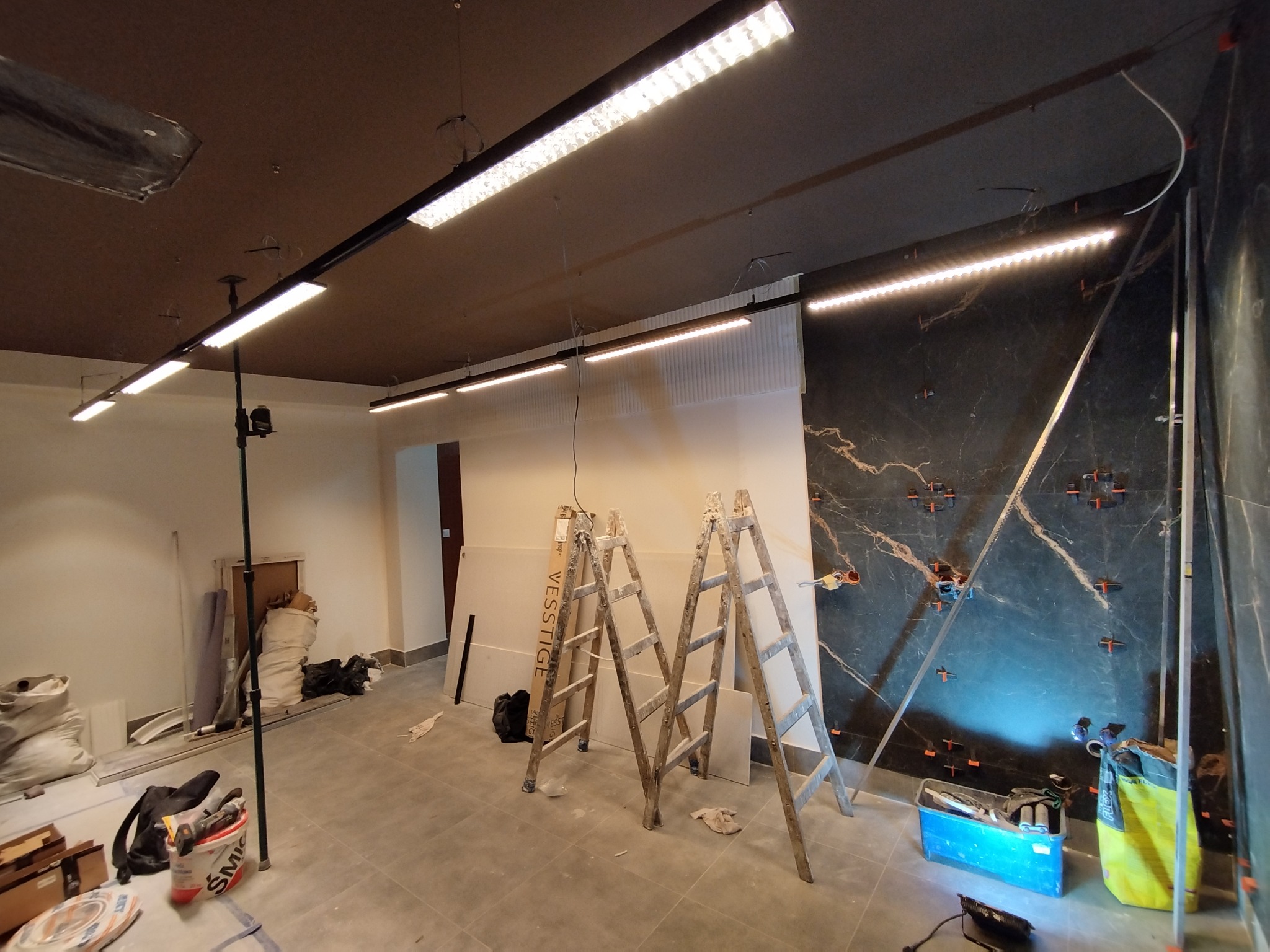 Montaż liniowego oświetlenia LED w pomieszczeniu z marmurową ścianą, widoczne drabiny i narzędzia.