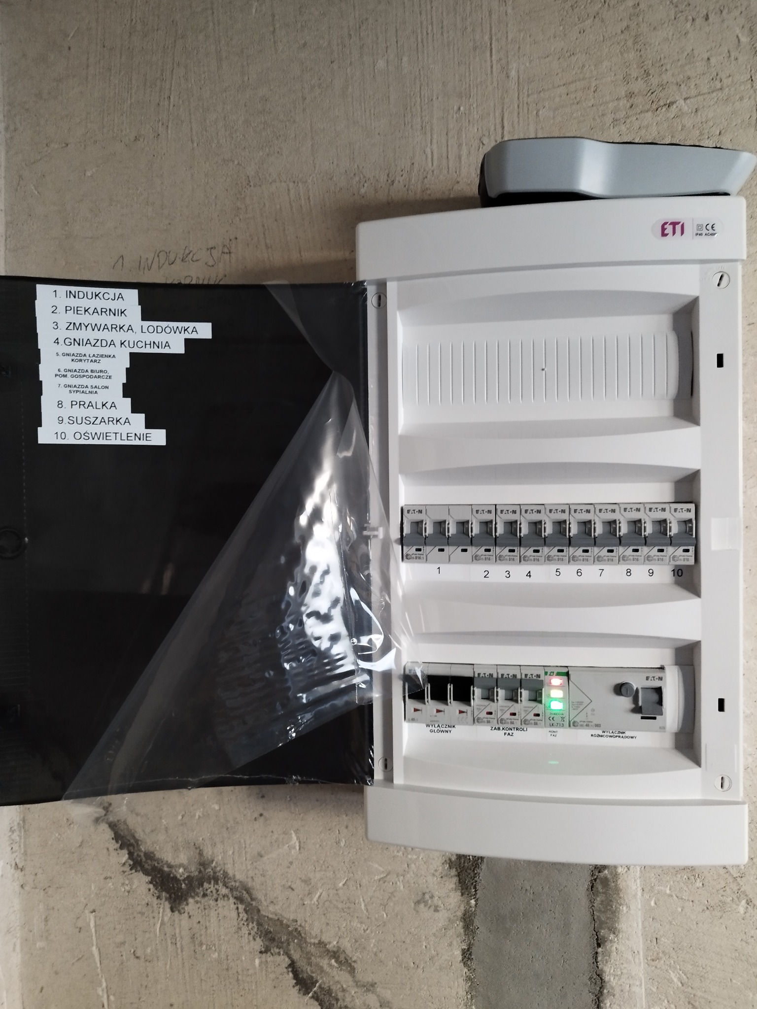 Otwarta skrzynka rozdzielcza z opisem obwodów (indukcja, piekarnik, zmywarka, gniazda kuchenne, łazienkowe, biuro, salon, pralka, suszarka, oświetlenie) i widocznymi bezpiecznikami, zamontowana...