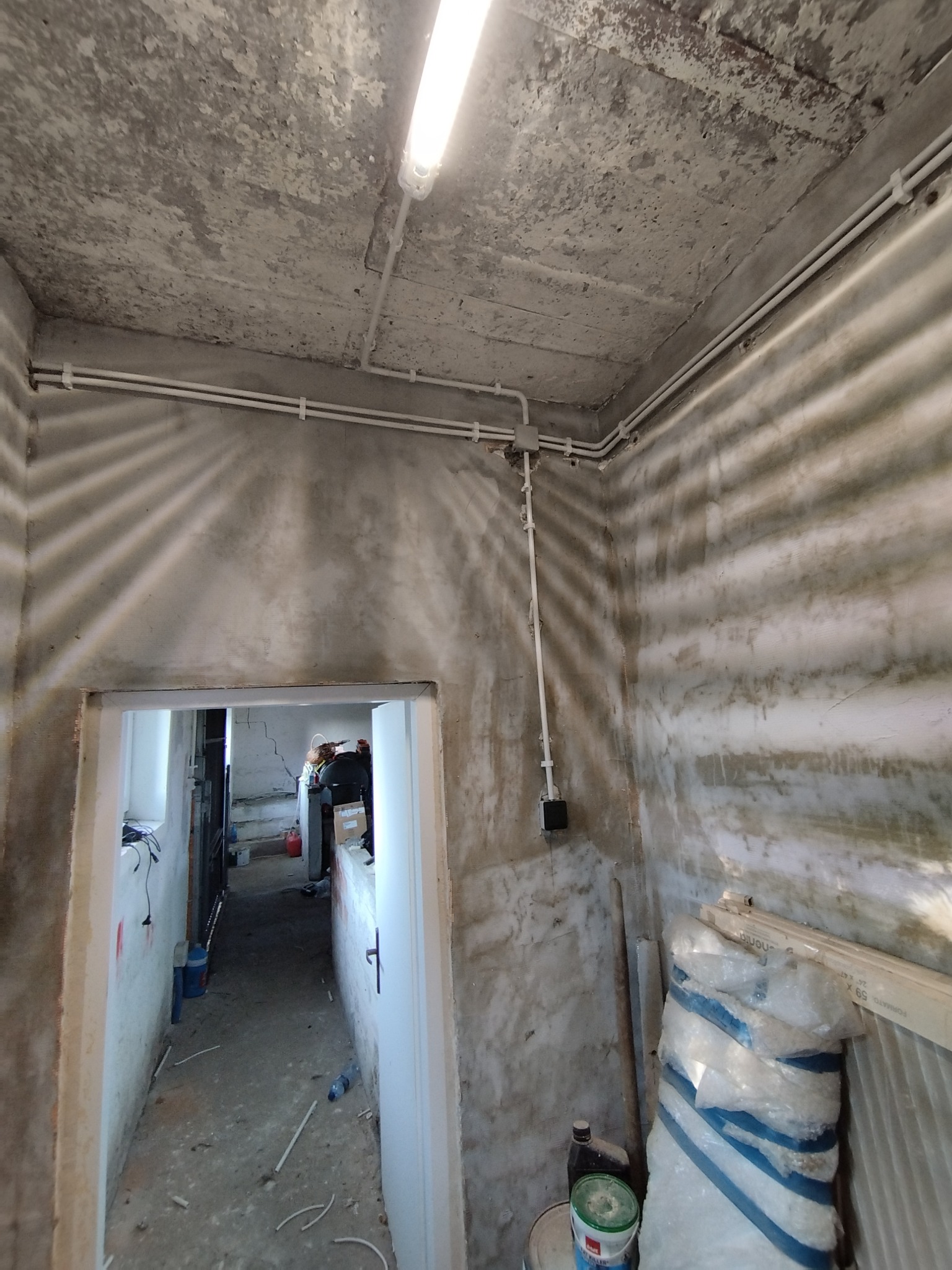 Surowy pokój z widocznymi rurami instalacji elektrycznej pod sufitem, lampą jarzeniową, betonową ścianą i podłogą w trakcie remontu, widok na dalsze pomieszczenia przez otwarte drzwi.
