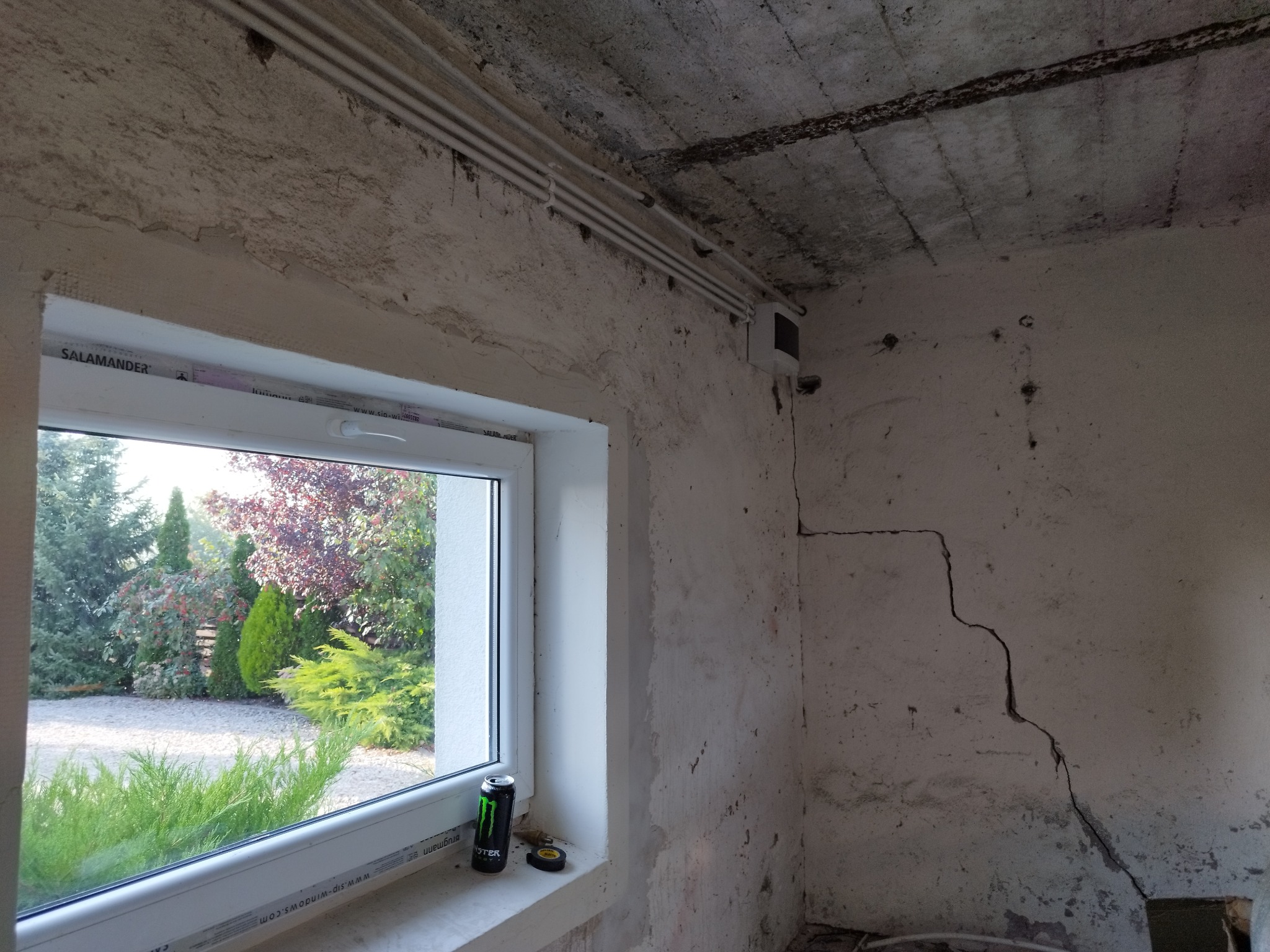 Surowe wnętrze z widocznymi rurami na ścianie, pęknięciem na rogu i oknem z widokiem na ogród. Na parapecie puszka napoju energetycznego i rolka taśmy izolacyjnej.