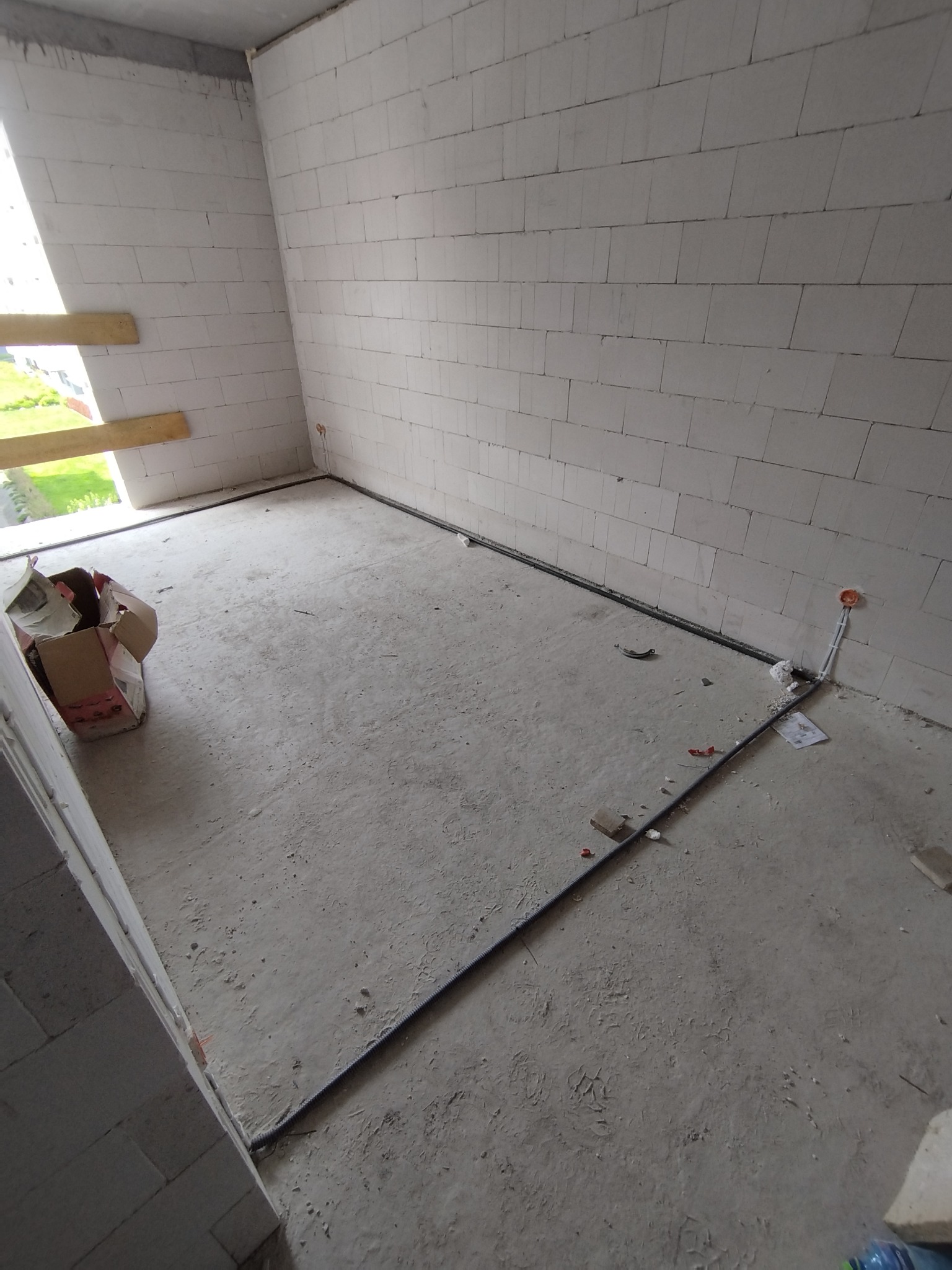 Surowy stan wnętrza z widocznymi przewodami elektrycznymi w peszlu ułożonymi wzdłuż ściany oraz puszką instalacyjną w ścianie, betonowa podłoga z resztkami materiałów budowlanych.
