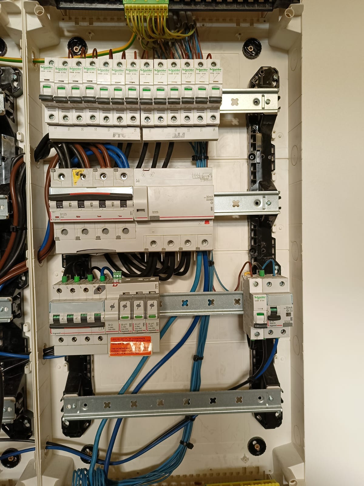 Otwarta skrzynka rozdzielcza z zamontowanymi bezpiecznikami i przewodami w różnych kolorach, widoczne elementy montażowe i naklejki informacyjne.