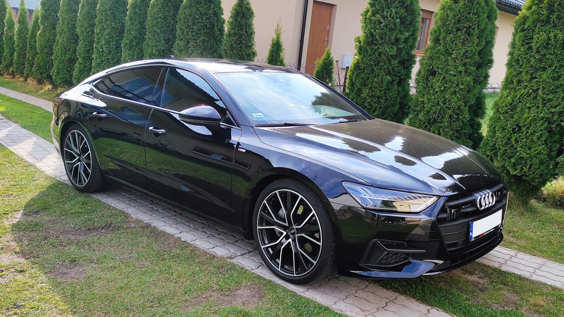 Czarna Audi A7 C8 po przyciemnieniu szyb, zaparkowana na kostce brukowej przed tujami, widoczny efekt przyciemnienia przednich szyb.