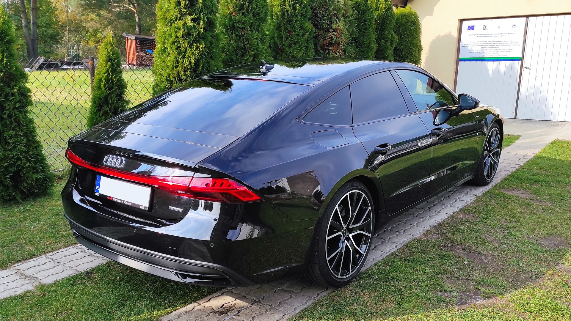 Czarny Audi A7 C8 po zabiegu zmiany stopnia przepuszczalności światła w szybach, zaparkowany na trawniku przed budynkiem z jasną elewacją i tujami.