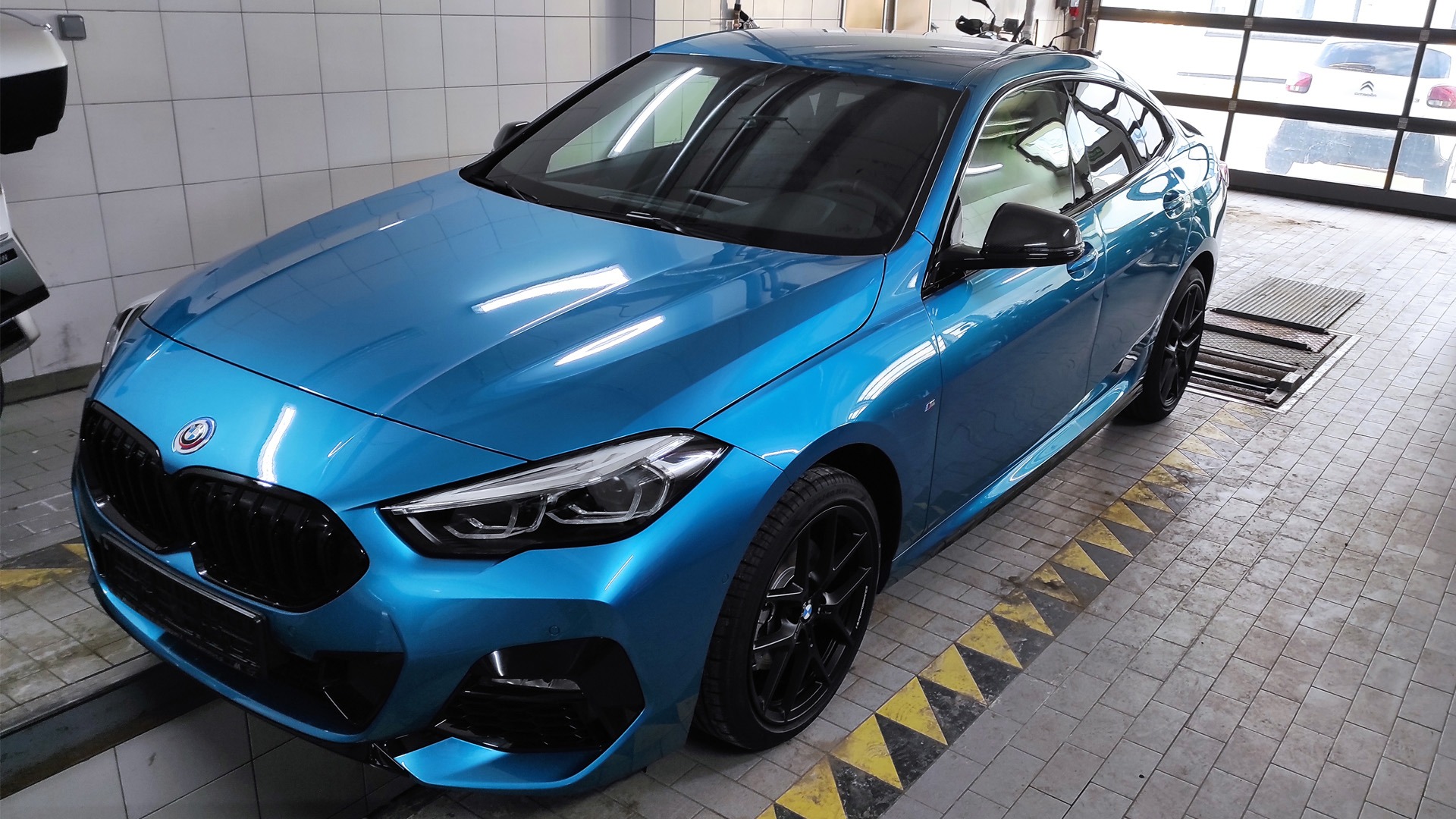 Błękitne BMW 2 Gran Coupe po zabiegu przyciemniania szyb, zaparkowane w warsztacie z kratką odpływową w podłodze i białymi płytkami na ścianach.
