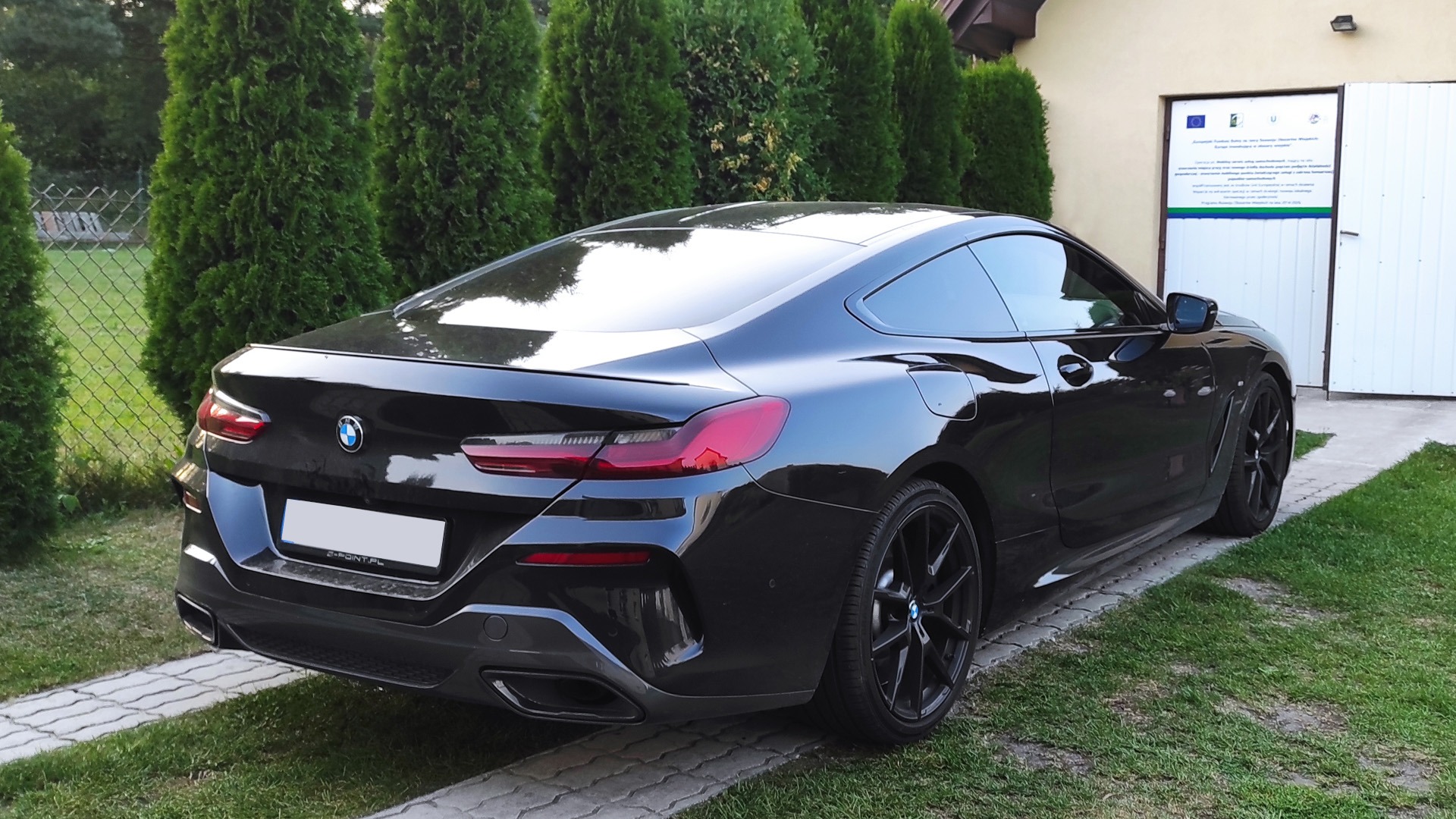 Czarny BMW Seria 8 Coupe po zabiegu przyciemniania szyb, zaparkowany na trawniku przed budynkiem z białymi drzwiami i tablicą informacyjną. Widok z tyłu, z wyraźnie widocznymi ciemnymi szybami...