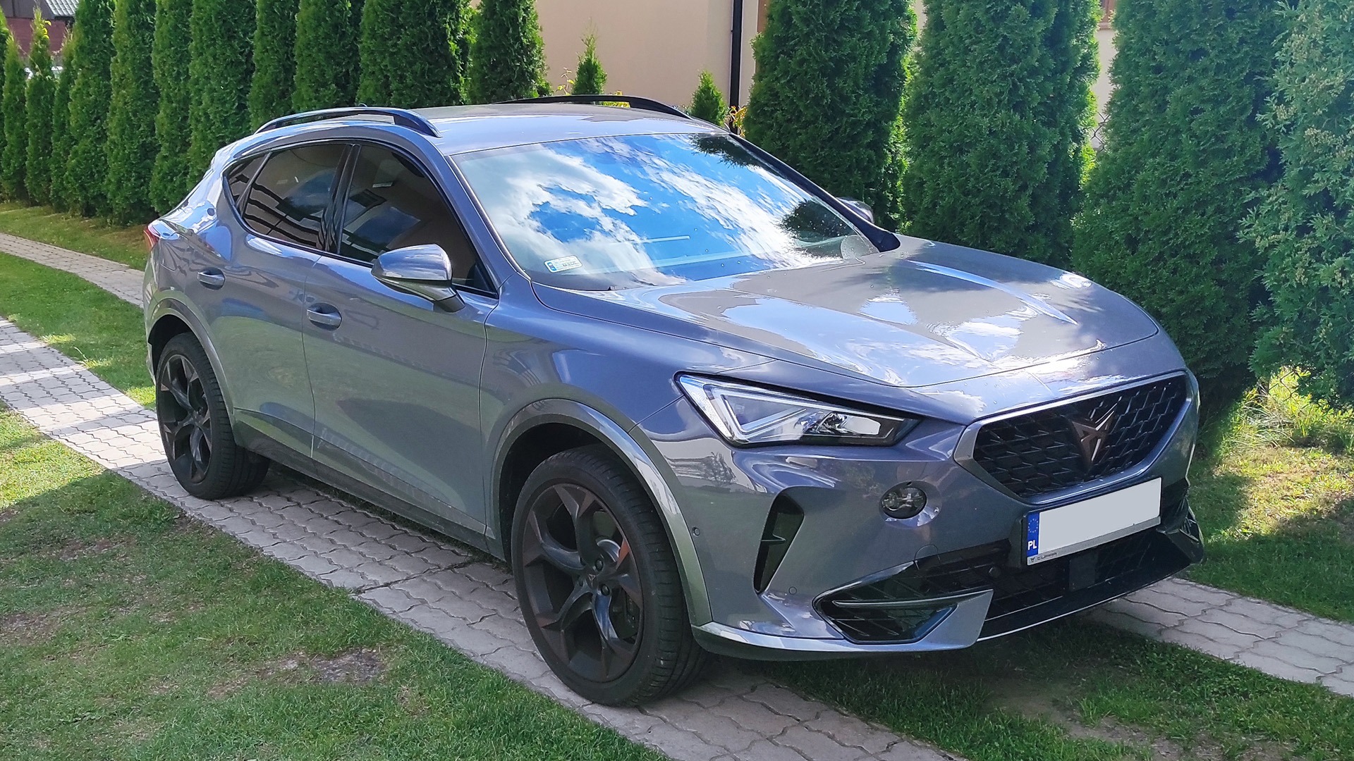 Szary samochód Cupra Formentor VZ z przyciemnionymi szybami, zaparkowany na podjeździe z kostki, z widocznym odbiciem chmur na przedniej szybie i delikatnie przyciemnionymi szybami bocznymi.