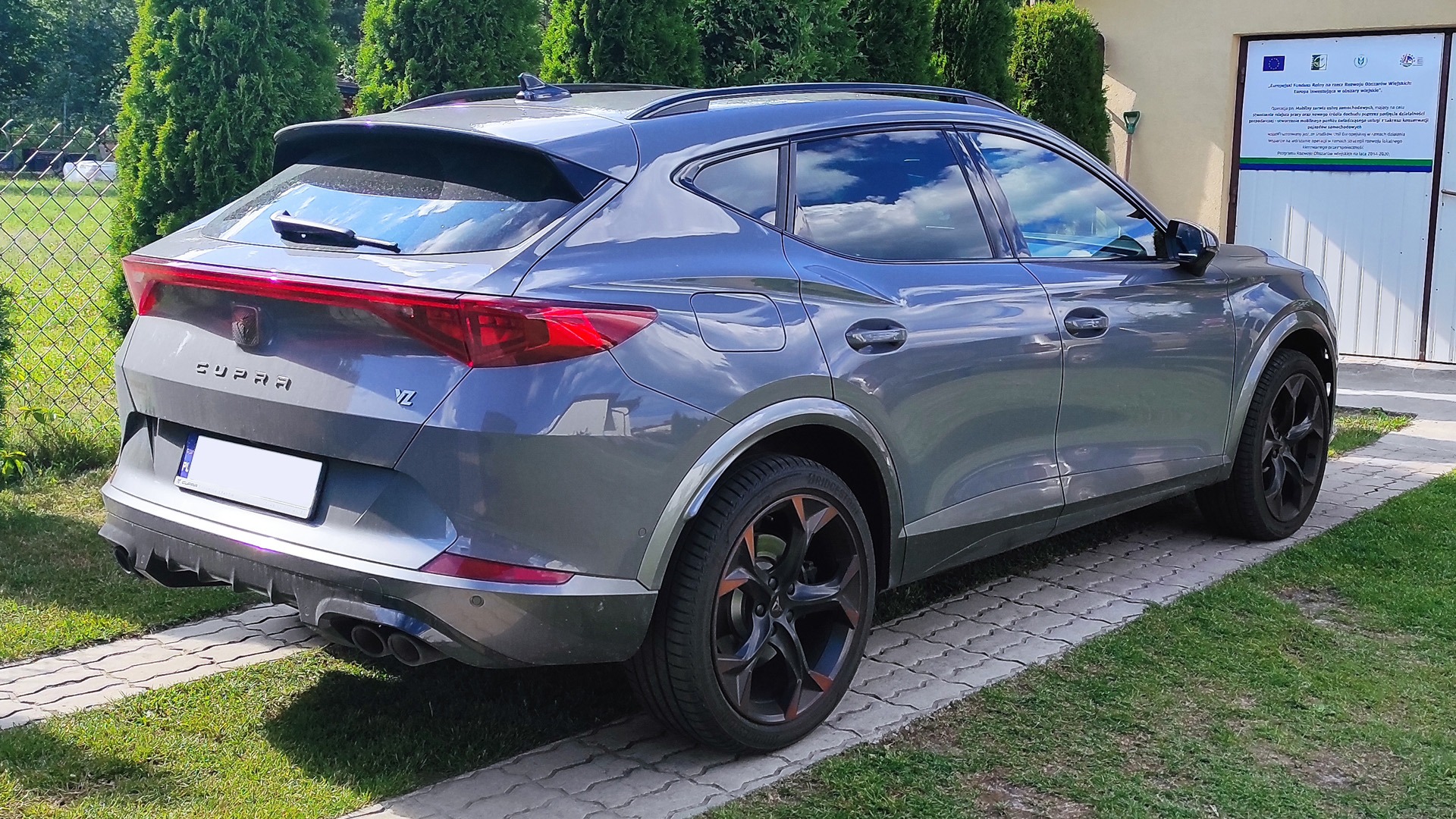 Szary samochód Cupra Formentor VZ z przyciemnionymi szybami, widok z tyłu, na tle trawnika i budynku z tablicą informacyjną.