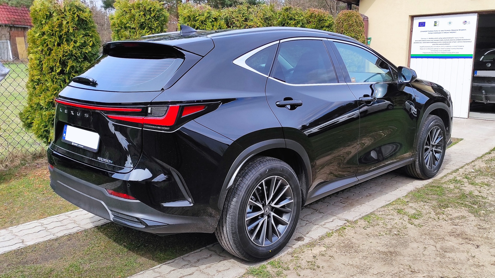 Czarny Lexus NX 350h po zabiegu zaciemniania szyb, widok z tyłu, przyciemnione szyby tylne, jasne otoczenie z zielenią i budynkiem w tle.
