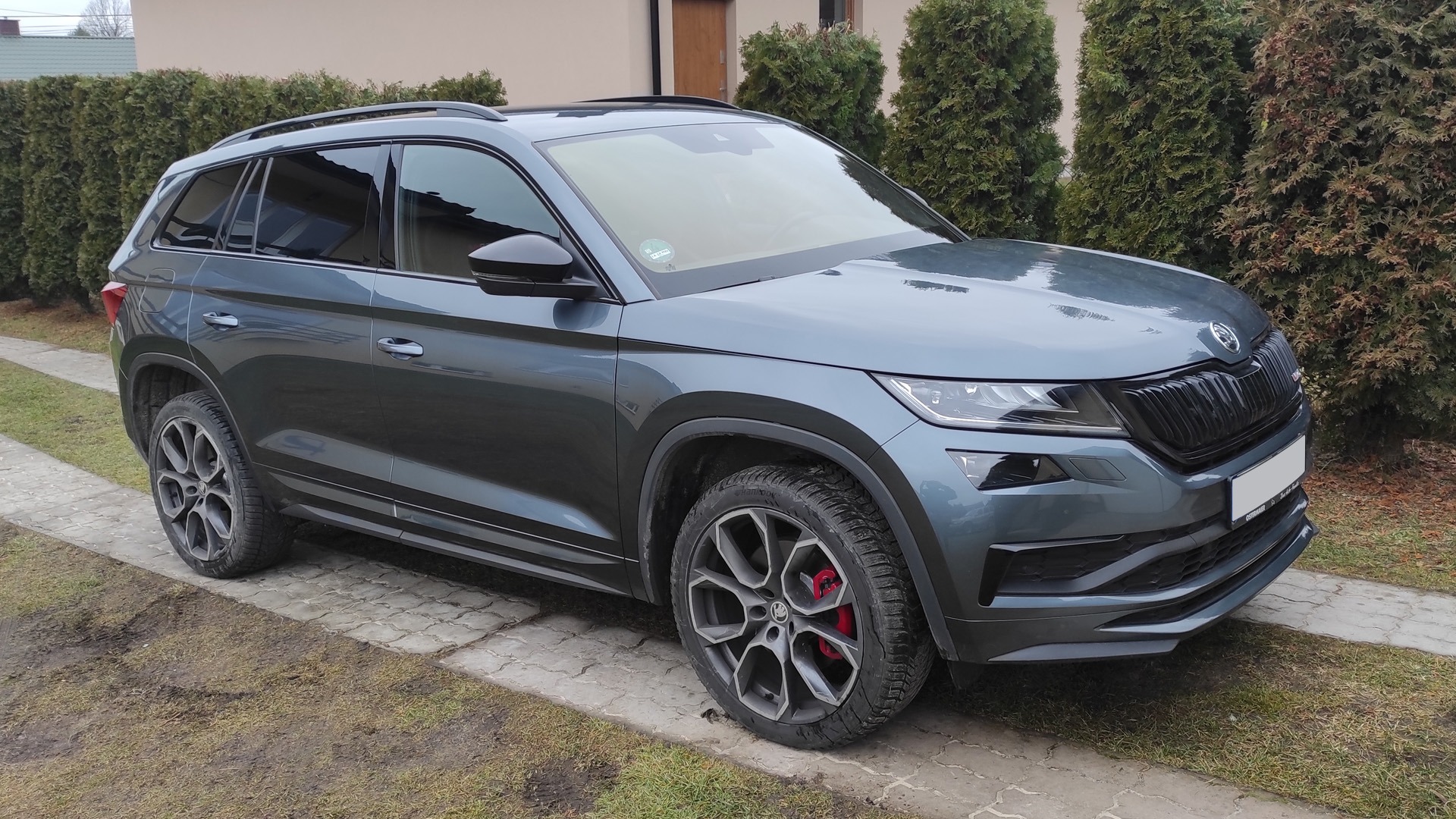 Szara Skoda Kodiaq VRS z przyciemnionymi tylnymi szybami, folia ceramiczna, widoczne czerwone zaciski hamulcowe, stojąca na kostce brukowej przed żywopłotem.