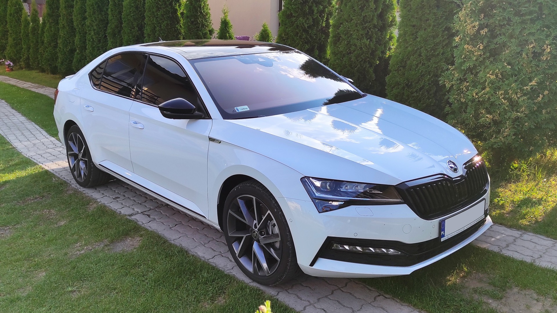 Biała Skoda Superb III po zabiegu przyciemniania szyb, zaparkowana na posesji w Stąporkowie. Widoczne ciemne szyby, szczególnie tylne, kontrastujące z jasnym lakierem karoserii. Ujęcie z przodu...