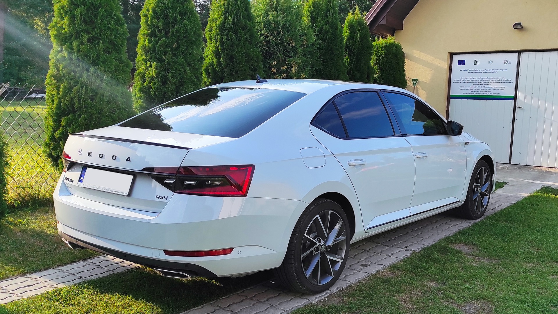 Biała Skoda Superb III z przyciemnionymi szybami, zaparkowana na kostce brukowej przed budynkiem z zielonymi tujami w tle. Widoczny efekt przyciemnienia szyb tylnych.