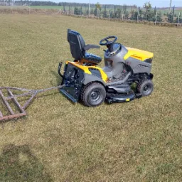 Żółto-szary traktorek do koszenia trawy z podczepioną broną na trawniku, w tle drzewa i ogrodzenie.
