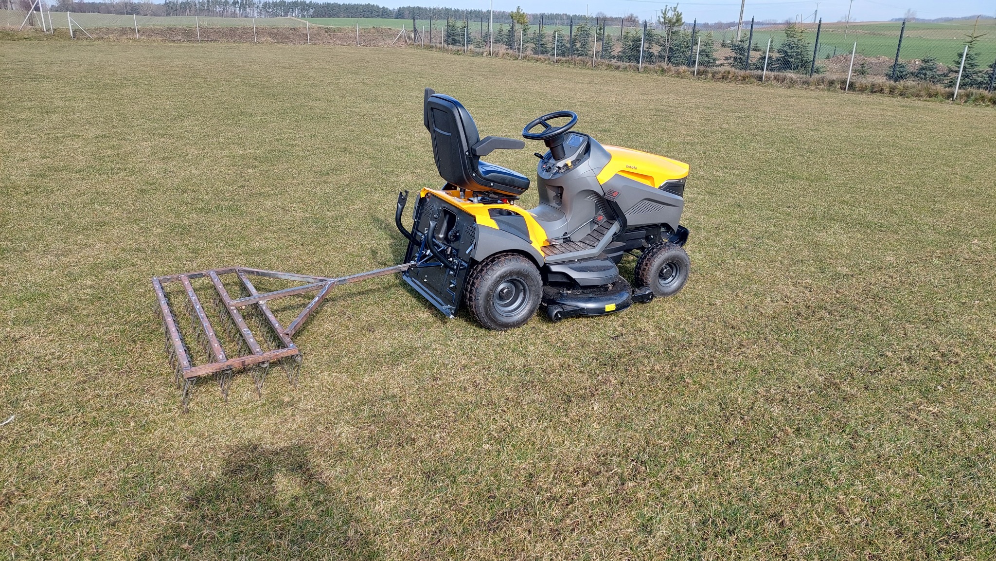 Żółto-szary traktorek do koszenia trawy z podczepioną broną na trawniku, w tle drzewa i ogrodzenie.
