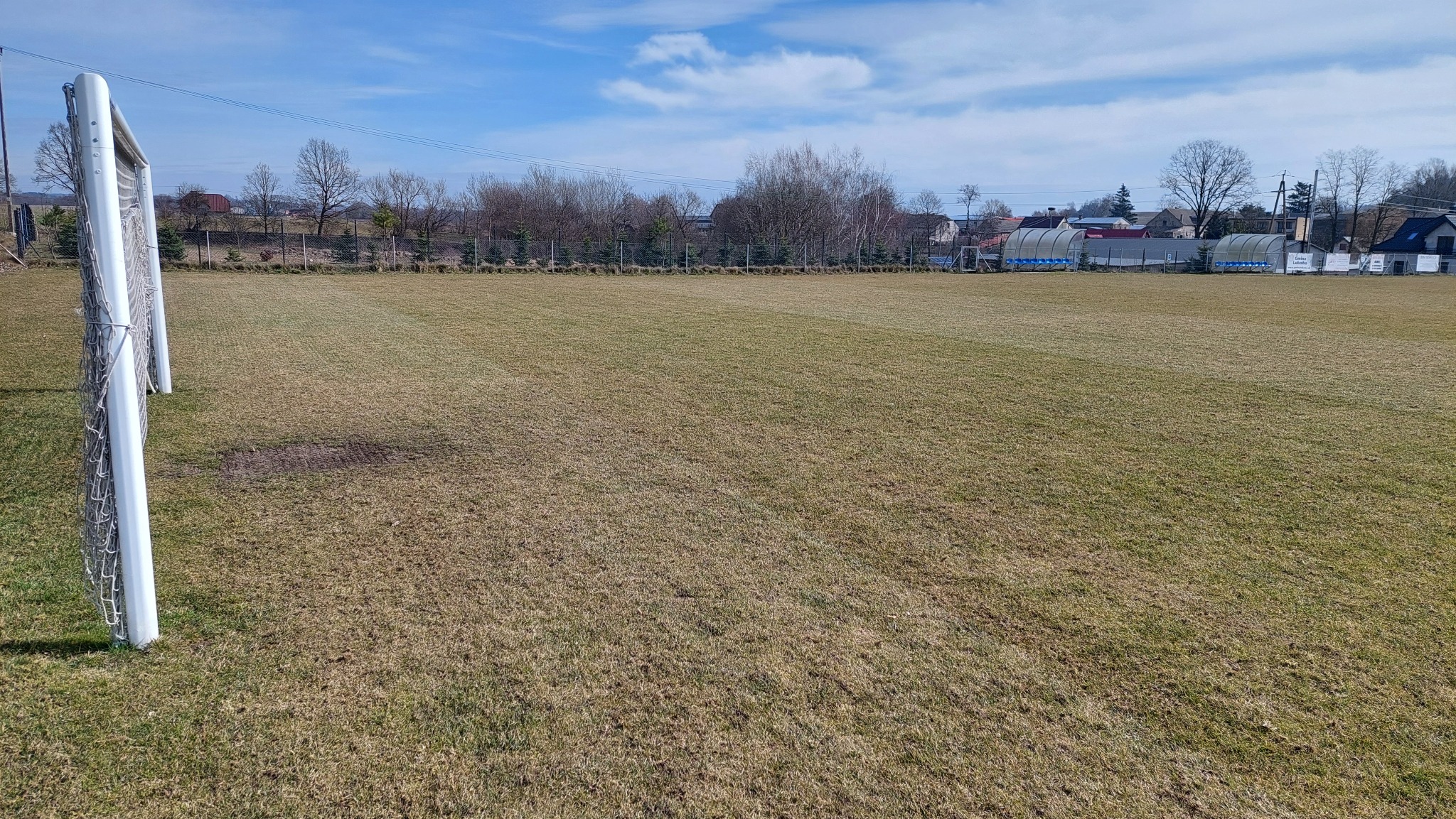 Widok na boisko sportowe z bramką, trawa częściowo wyschnięta, w tle zabudowania wiejskie i drzewa bez liści pod błękitnym niebem.