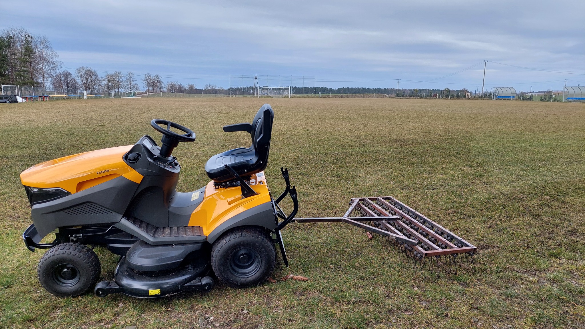 Żółty traktorek do koszenia trawy z doczepioną broną na boisku sportowym, w tle widoczne bramki i drzewa.