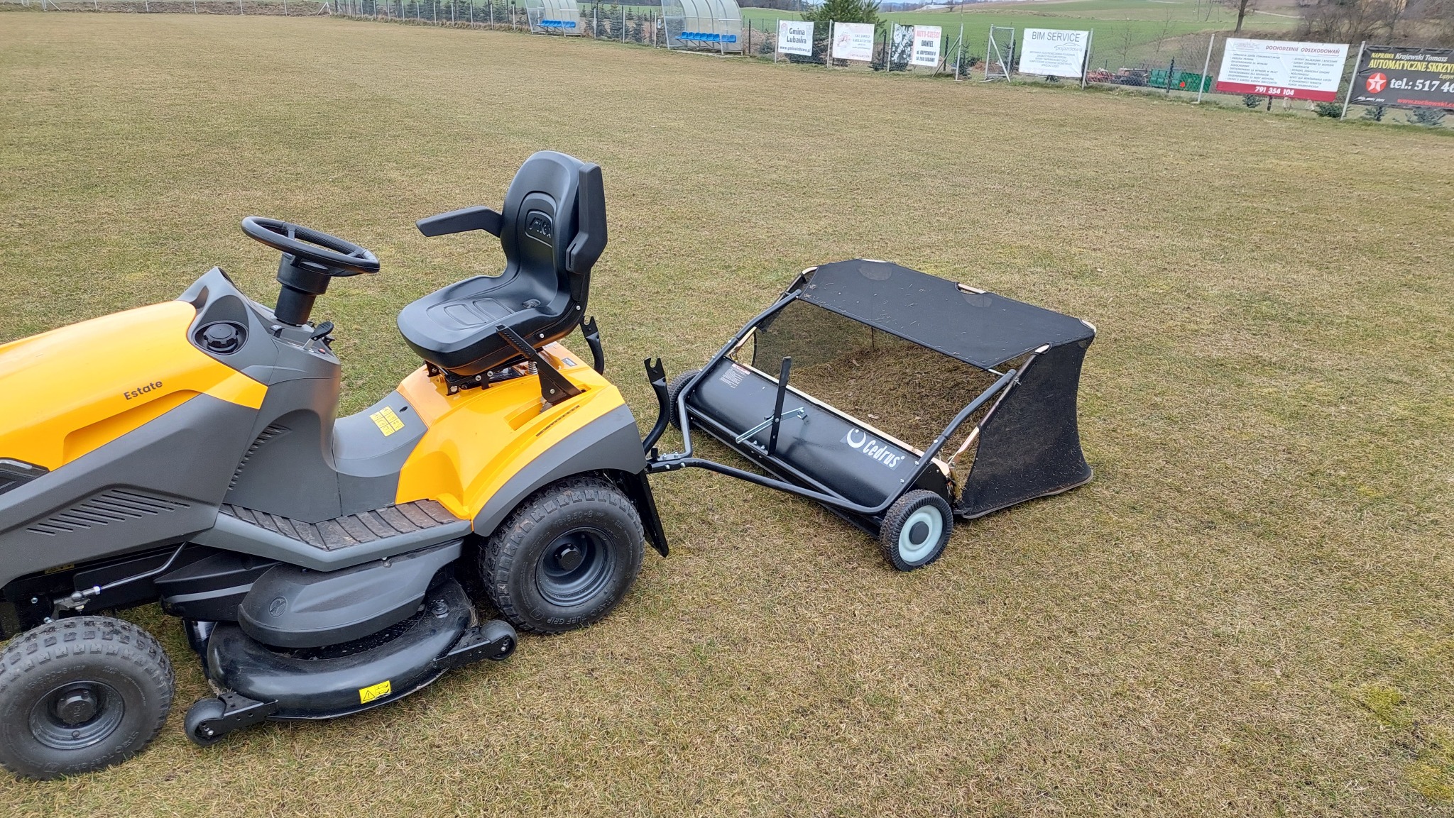 Żółto-szary traktorek ogrodowy z doczepionym koszem na trawę, stojący na trawniku. W tle widoczne ogrodzenie i reklamy.