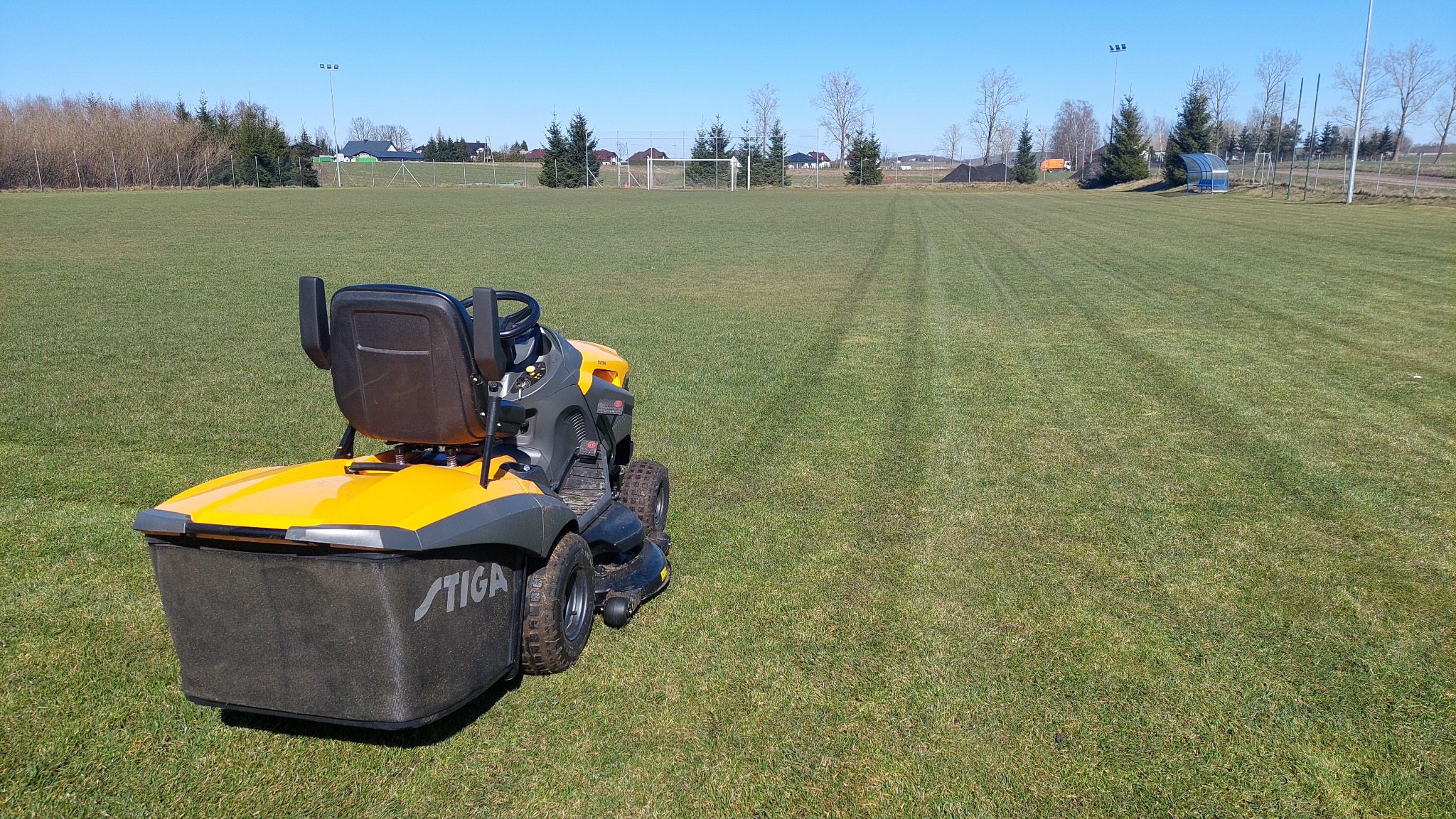 Żółto-szara kosiarka samojezdna Stiga na przystrzyżonym trawniku boiska sportowego, widoczne pasy skoszonej trawy w kierunku bramek.