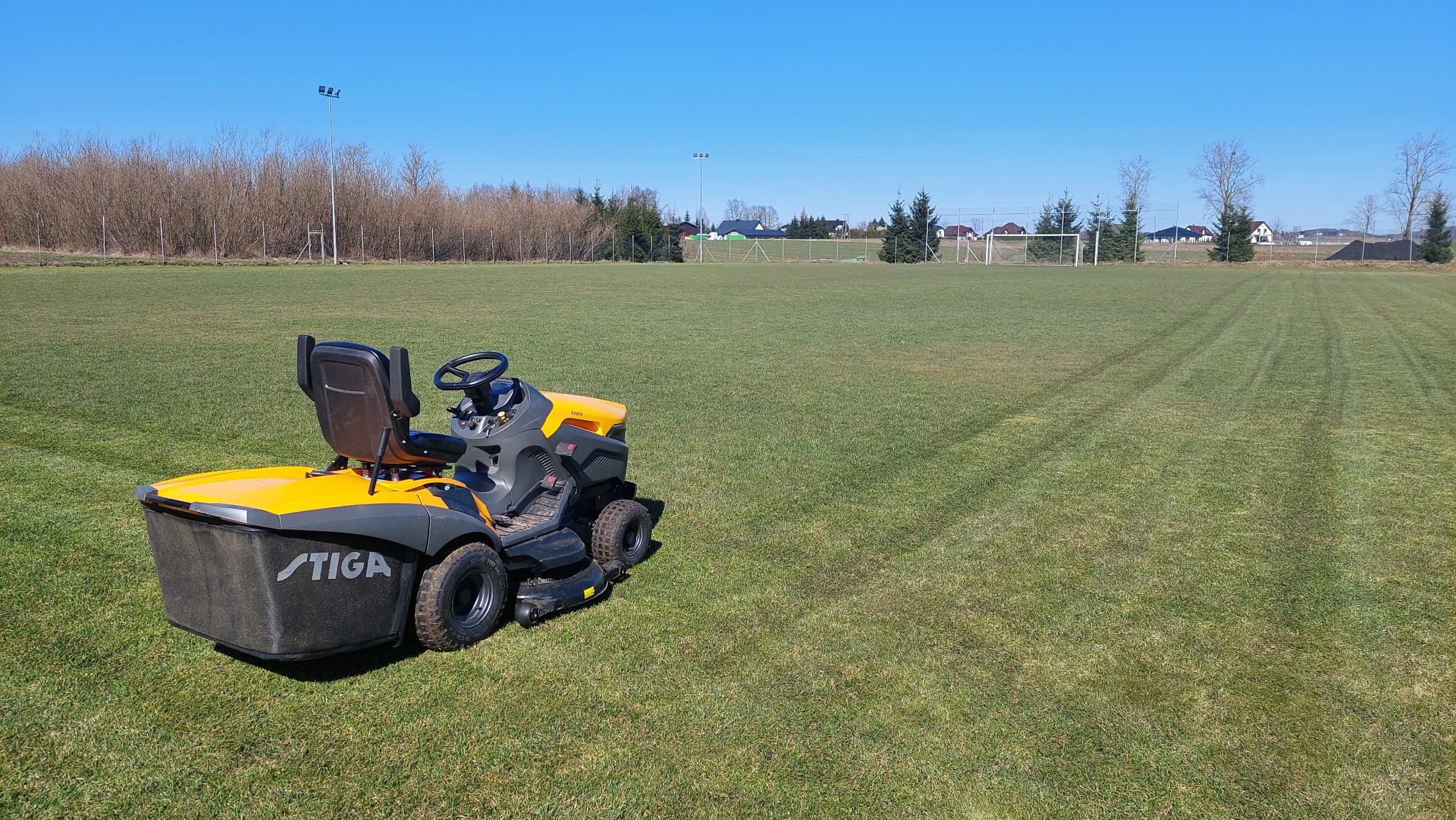 Żółto-szara kosiarka samojezdna marki Stiga na tle przystrzyżonej murawy boiska sportowego z widocznymi pasami, w oddali bramka i drzewa, błękitne niebo.