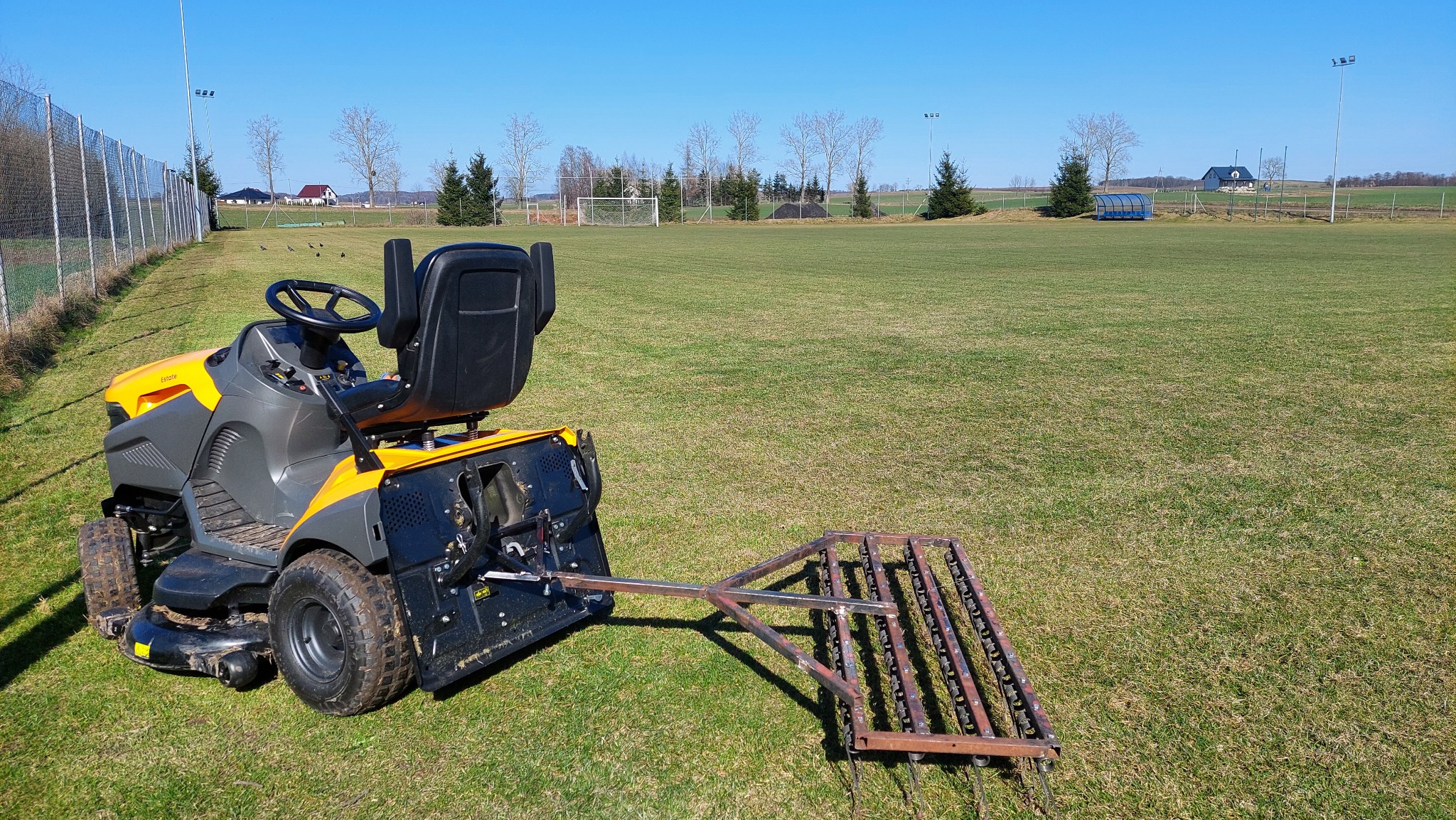 Żółto-szara kosiarka traktorowa z zamontowanym aeratorem na tle boiska sportowego w słoneczny dzień.