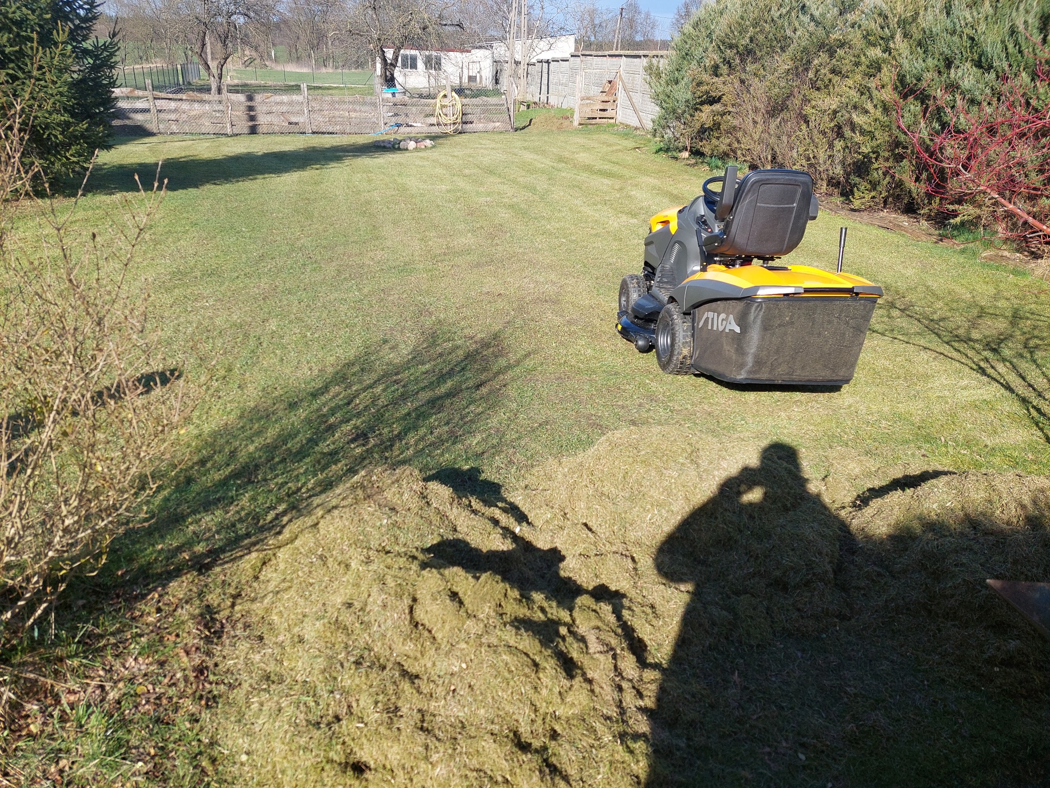 Żółto-szara kosiarka traktorowa STIGA na trawniku z widocznymi śladami koszenia i stertą skoszonej trawy w cieniu, zarys sylwetki fotografa.