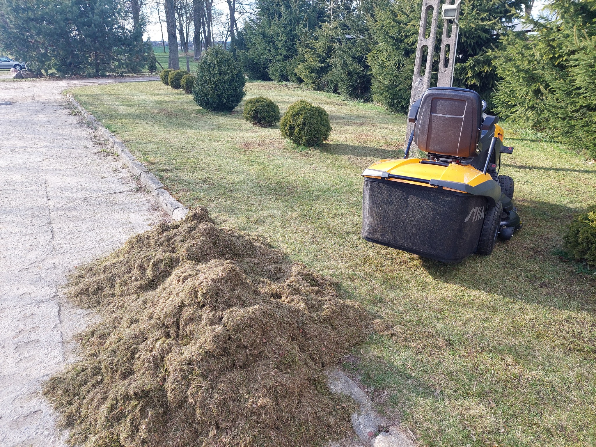 Żółto-czarna kosiarka samojezdna marki Stiga stoi na trawniku obok dużej pryzmy resztek po wertykulacji trawnika, w tle tuje i drzewa.