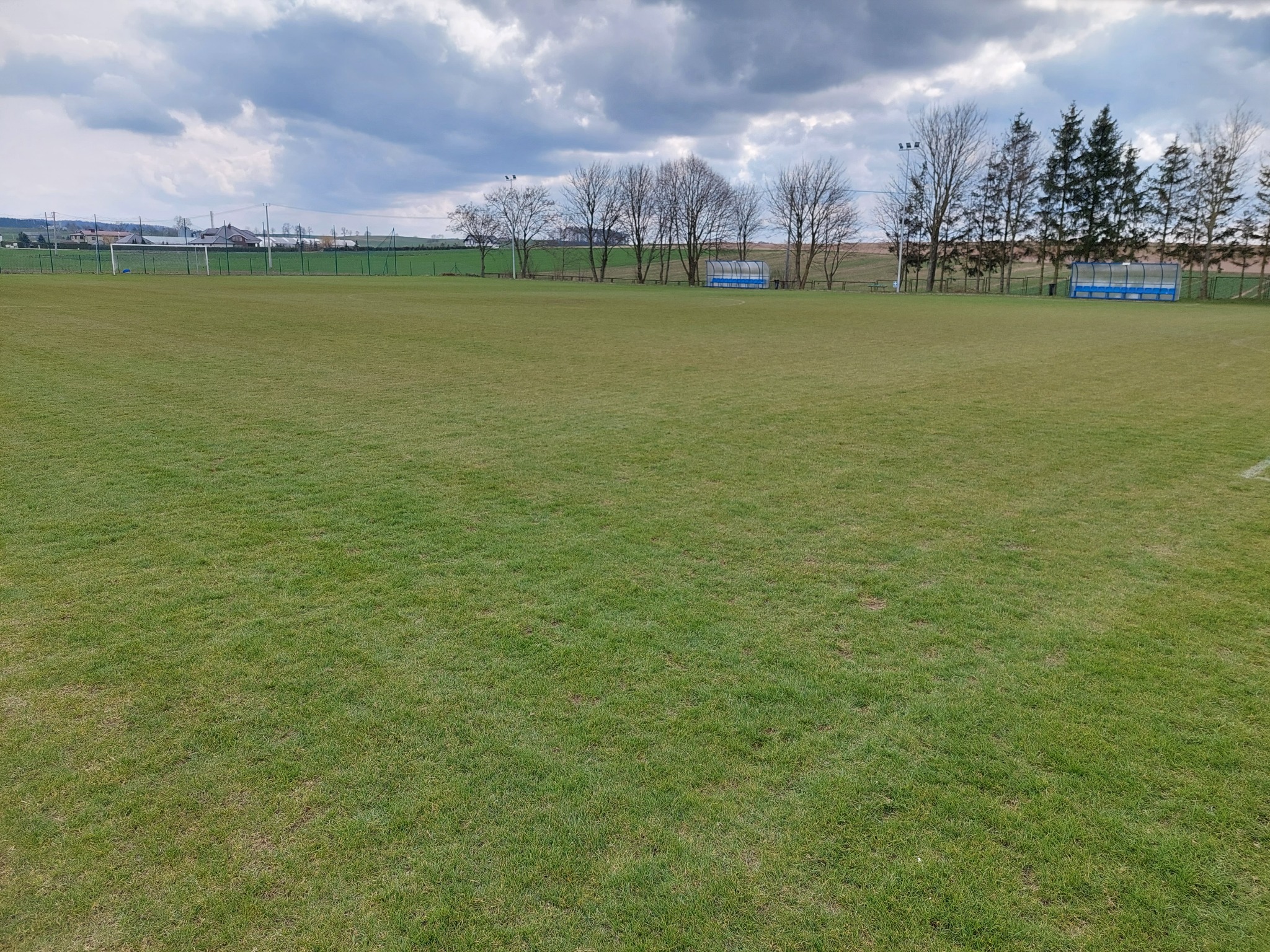 Rozległe boisko sportowe z równo przystrzyżoną trawą, widoczne bramki i zadaszone ławki dla zawodników w tle, pochmurne niebo.