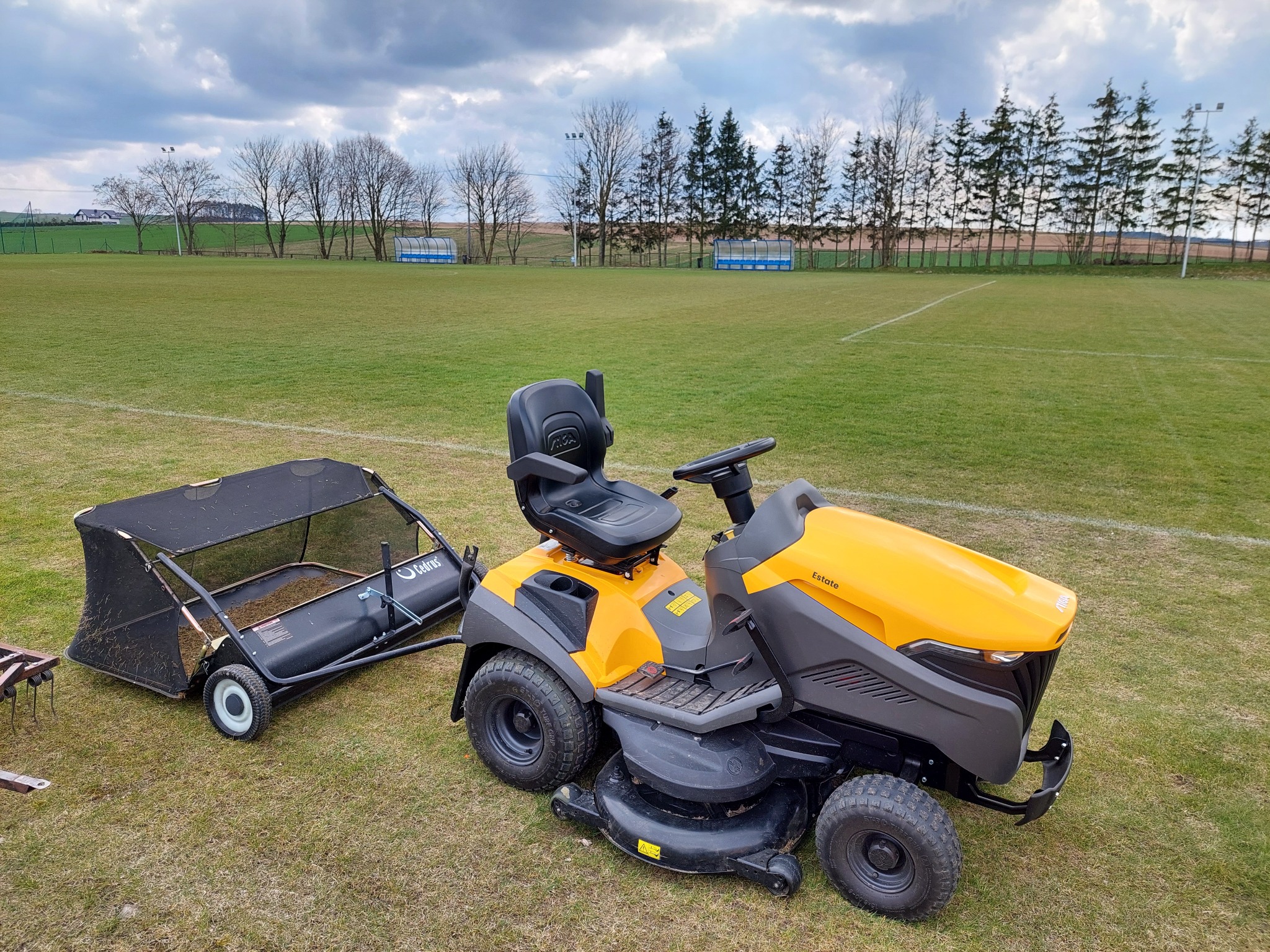 Żółto-czarny traktorek do koszenia trawy z doczepionym koszem na trawę stoi na zielonym boisku sportowym z widocznymi liniami, w tle drzewa i pochmurne niebo.