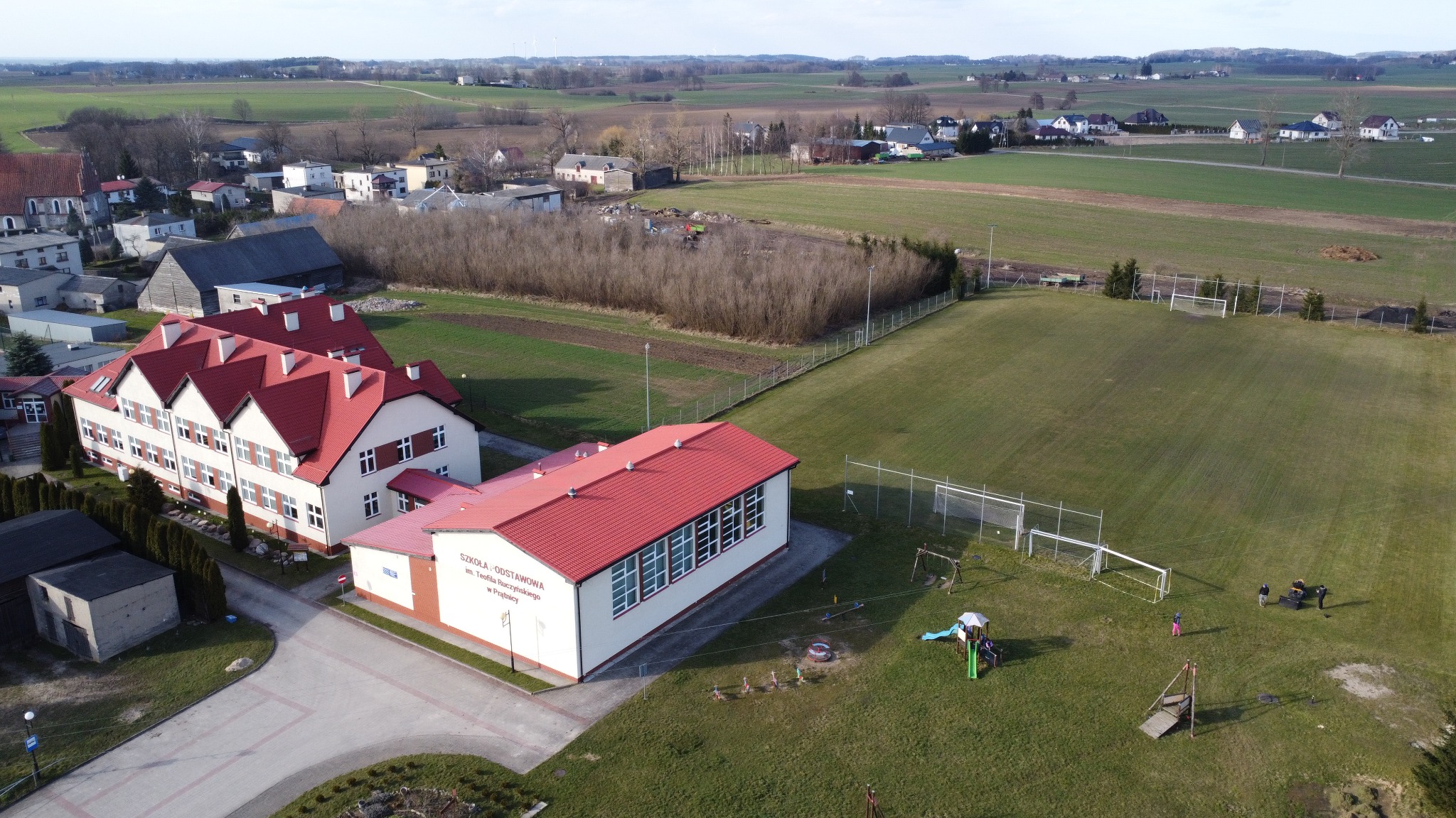 Widok z lotu ptaka na szkołę podstawową z czerwonym dachem i rozległym terenem zielonym, w tle pola uprawne i zabudowania wiejskie, widoczne boisko sportowe i plac zabaw.