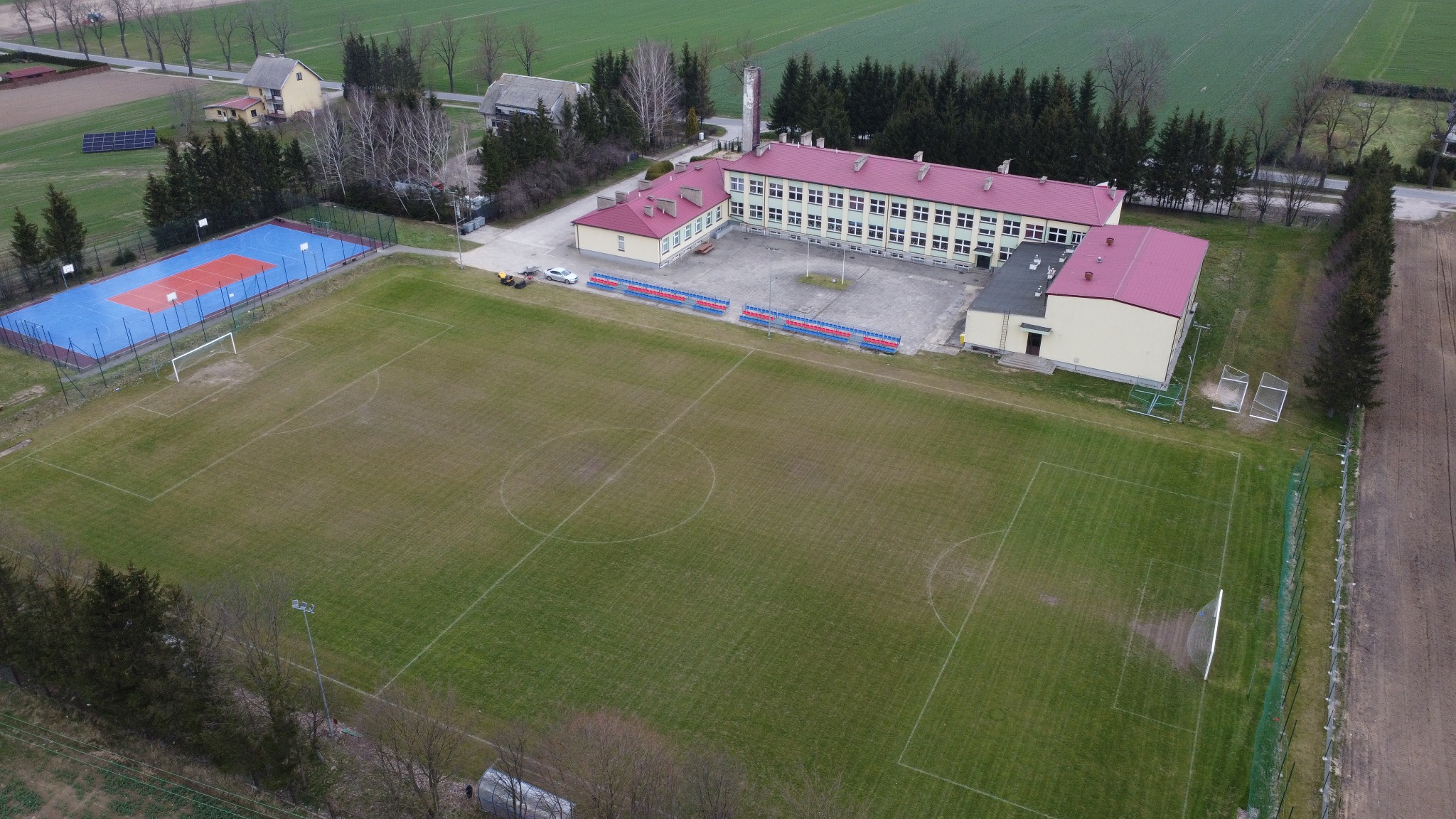Widok z drona na boisko szkolne z zieloną trawą, budynek szkoły z czerwonym dachem, boisko do koszykówki i siatkówki z niebieską nawierzchnią oraz otaczające drzewa i pola.