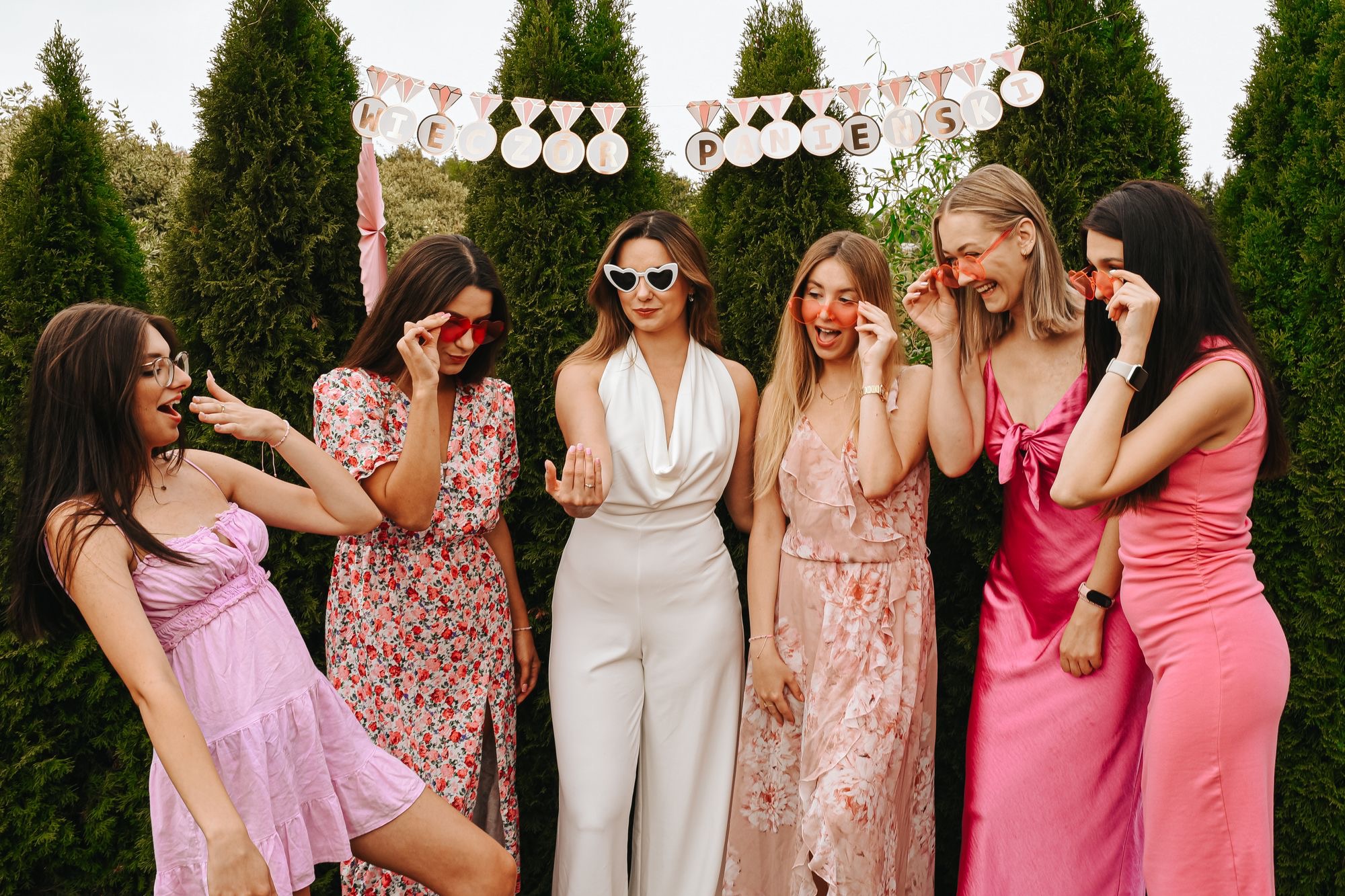 Grupa sześciu kobiet w różowych i białych strojach pozuje na tle zielonych drzew, dekoracja z napisem 'Wieczór Panieński' w tle, okulary w kształcie serc.