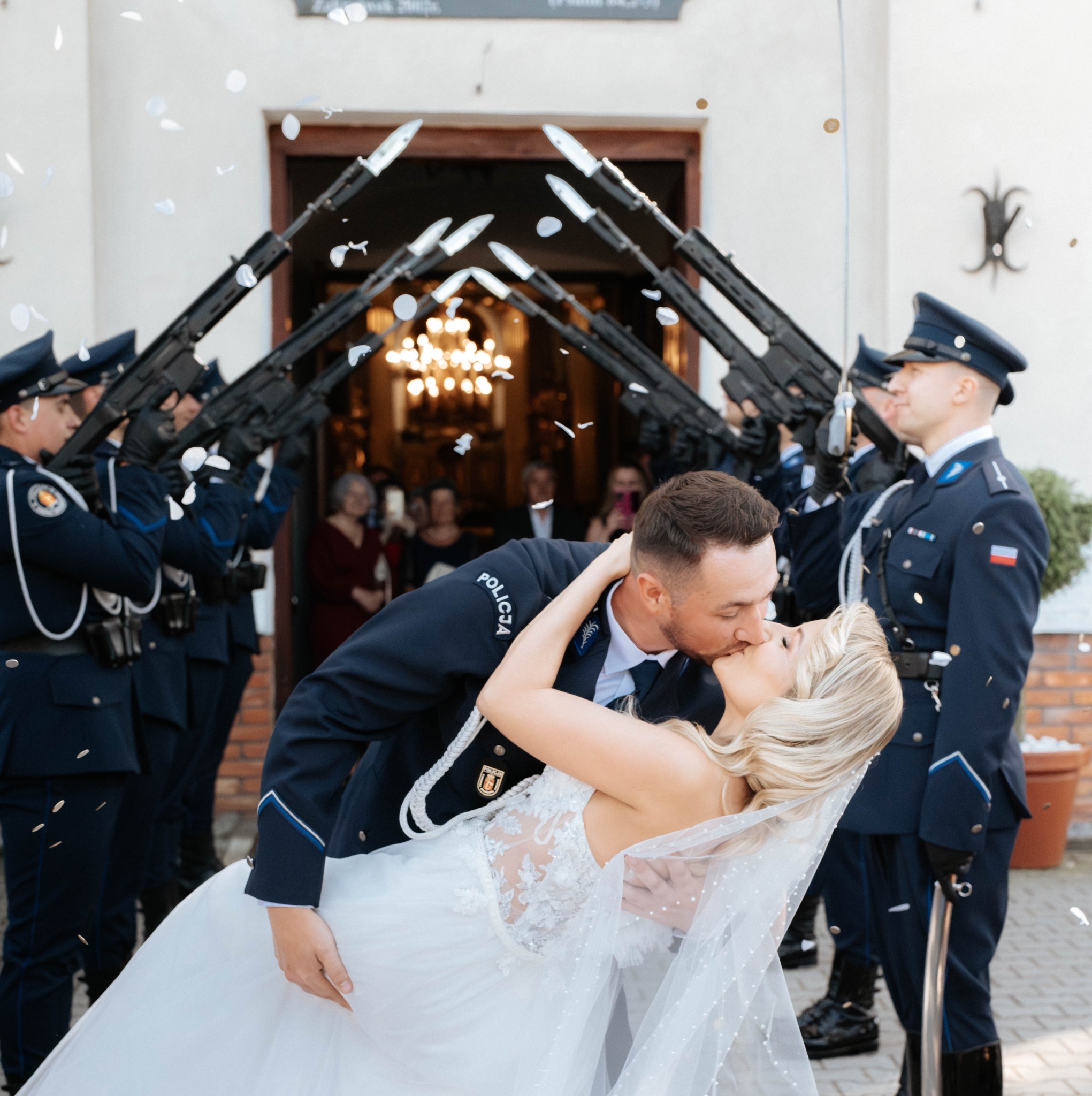 Pocałunek pary młodej w mundurze policyjnym pod szpalerem karabinów, konfetti w powietrzu. Nietypowe, uroczyste tło ślubne.