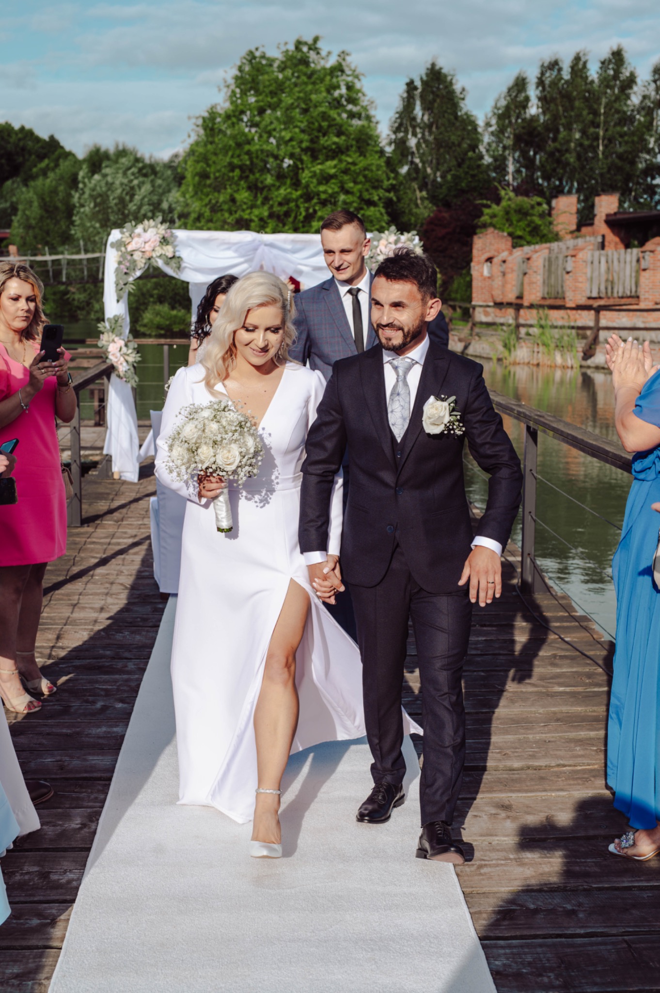 Para młoda, trzymając się za ręce, idzie po białym dywanie na drewnianym pomoście w otoczeniu gości i zieleni, w tle ruiny budynku.