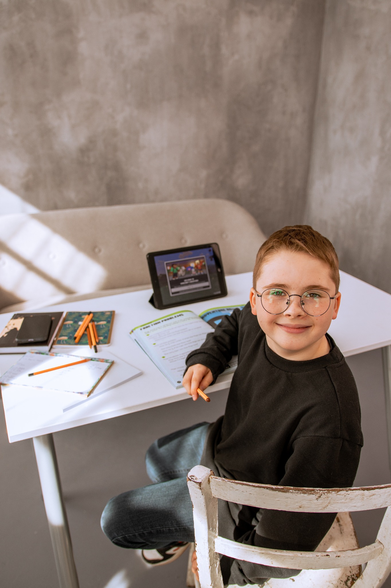 Uśmiechnięty chłopiec w okularach siedzi przy białym biurku z tabletem, zeszytami i podręcznikiem, trzymając ołówek w ręce.