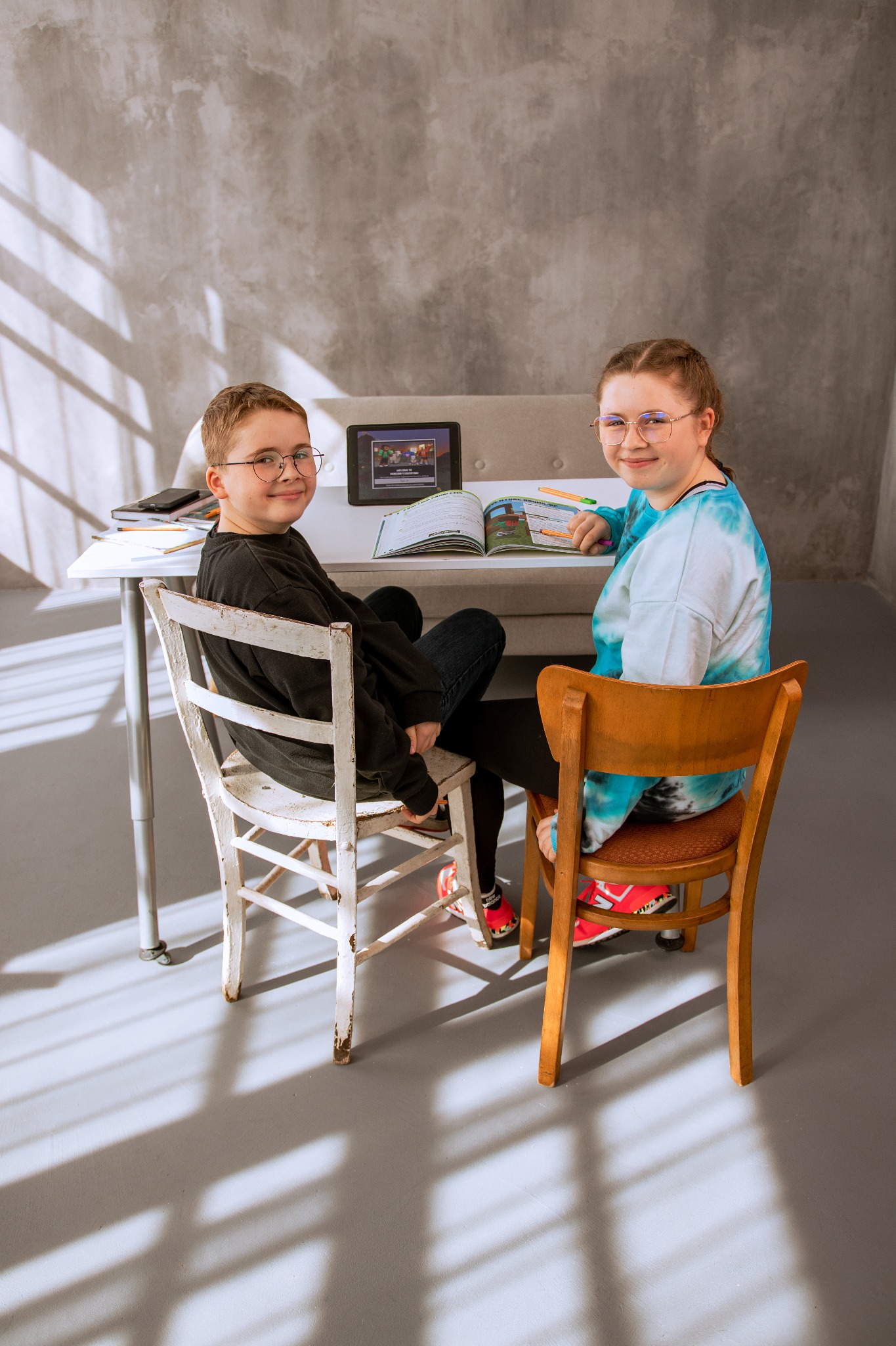 Dwoje uczniów siedzi przy biurku z otwartym podręcznikiem i tabletem z grą Minecraft na ekranie, oświetleni promieniami słońca rzucającymi cienie na podłogę i ścianę z szarą fakturą.
