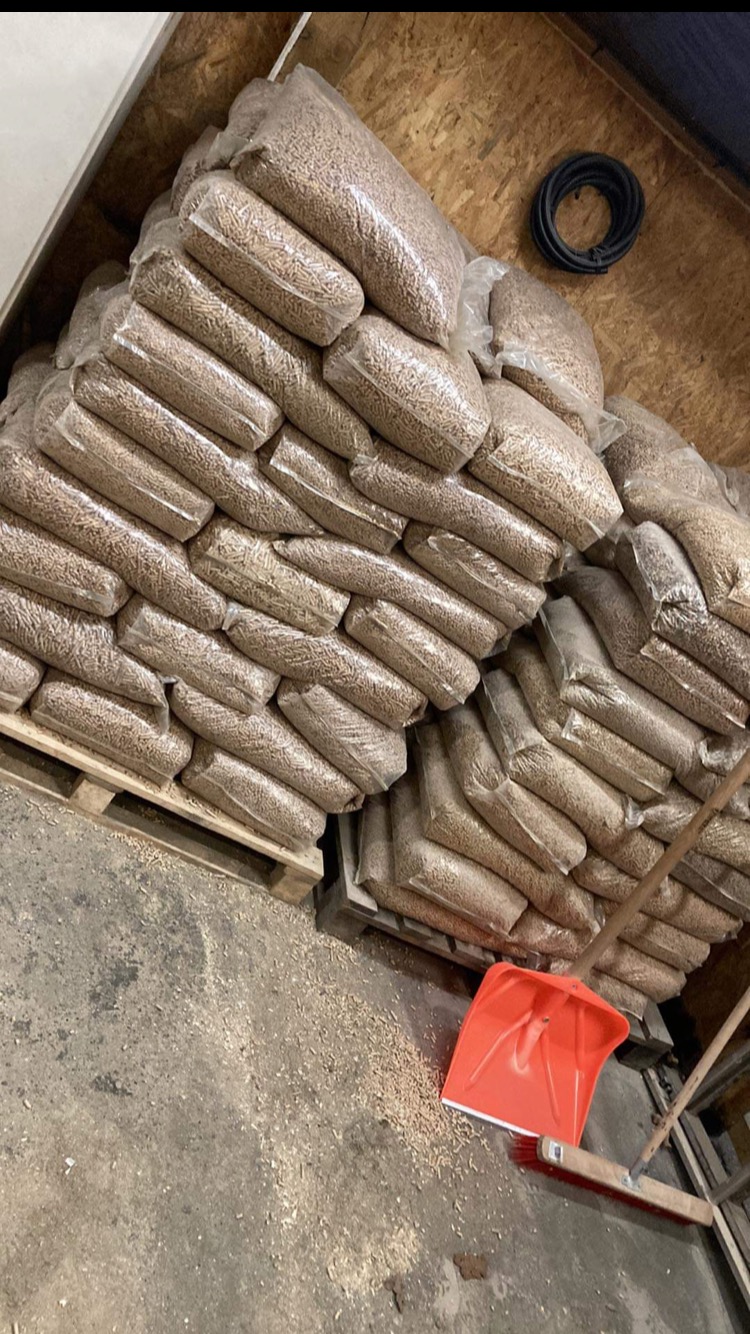 Stos worków z pelletem ułożonych na paletach w magazynie, widoczna czerwona łopata do zgarniania rozsypanego pelletu, podłoga z drobnymi trocinami.