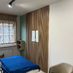 Nowoczesna sypialnia z drewnianą ścianą z lameli, oświetleniem punktowym na szynie, beżowym fotelem i niebieską narzutą na łóżku. Na ścianie zamontowany biały panel sterowania.