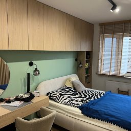 Face Front Konrad Olborski - Sypialnia z zabudową meblową z jasnego drewna nad łóżkiem, toaletka z okrągłym lustrem, ściana w kolorze pistacjowym, łóżko z narzutą w zebrę i niebieską kołdrą, drewniane żaluzje w oknie.