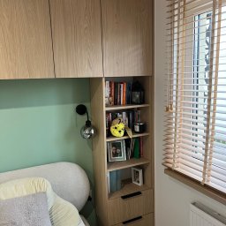 Face Front Konrad Olborski - Drewniana zabudowa nad łóżkiem z półkami i szafkami, oświetlona lampką ścienną, z widocznymi książkami i dekoracjami na półkach, oraz drewniane żaluzje w oknie.