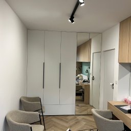 Face Front Konrad Olborski - Biała szafa z lustrzanymi drzwiami w pokoju z parkietem w jodełkę i dwoma fotelami, widok na biurko z okrągłym lustrem i oświetlenie sufitowe.