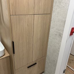 Face Front Konrad Olborski - Jasna, drewniana szafa na wymiar z czarnymi uchwytami, umiejscowiona w łazience z szarymi płytkami, widoczny fragment umywalki i drzwi z narzędziami montażowymi.