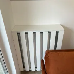 Biała, zabudowa grzejnika z pionowymi lamelami, dopasowana do wnęki pod skosem, obok stoi pomarańczowy fotel.