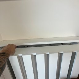 Face Front Konrad Olborski - Montaż białych, pionowych paneli dekoracyjnych na suficie, widoczna ręka z tatuażem przytrzymująca element mocujący.