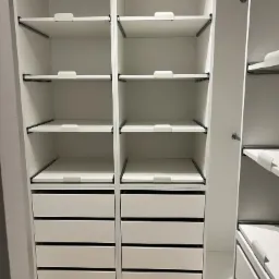 Wnętrze białej szafy wnękowej z szufladami i regulowanymi półkami, widoczne czarne metalowe wsporniki półek.