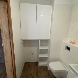 Face Front Konrad Olborski - Nowoczesna łazienka z białymi, wiszącymi szafkami, otwartymi półkami i podwieszaną toaletą na tle płytek imitujących drewno.