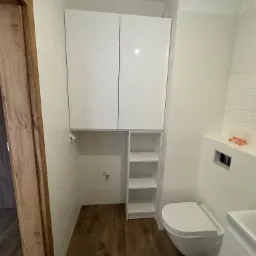 Nowoczesna łazienka z białymi, wiszącymi szafkami, otwartymi półkami i podwieszaną toaletą na tle płytek imitujących drewno.