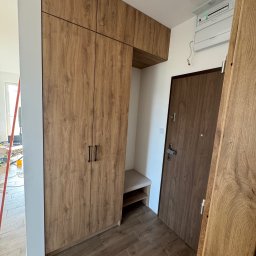 Face Front Konrad Olborski - Przedpokój z zabudową: szafa z drewnopodobnymi frontami i siedziskiem, dopasowana do drzwi wejściowych, widoczne niedokończone prace instalacyjne na suficie.