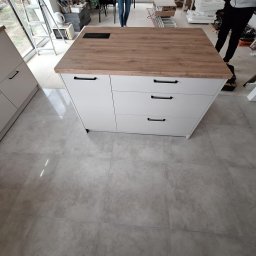 Face Front Konrad Olborski - Wyspa kuchenna z białymi frontami i blatem imitującym drewno, wyposażona w czarne, poziome uchwyty, widoczna w trakcie montażu w pomieszczeniu z szarą podłogą z płytek.