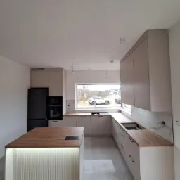 Nowoczesna kuchnia z wyspą, beżowe szafki z drewnianymi blatami, czarna lodówka i piekarnik, oświetlenie LED pod wyspą, okno z widokiem na zewnątrz.